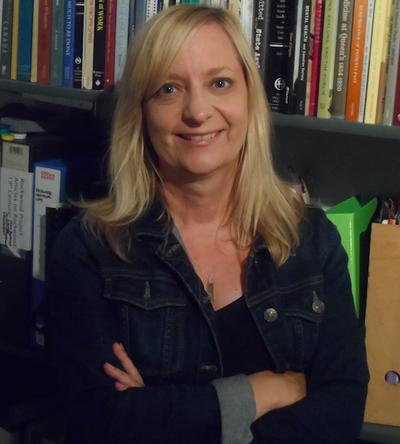 Kathy has long fair hair and is standing in front of bookshelves.