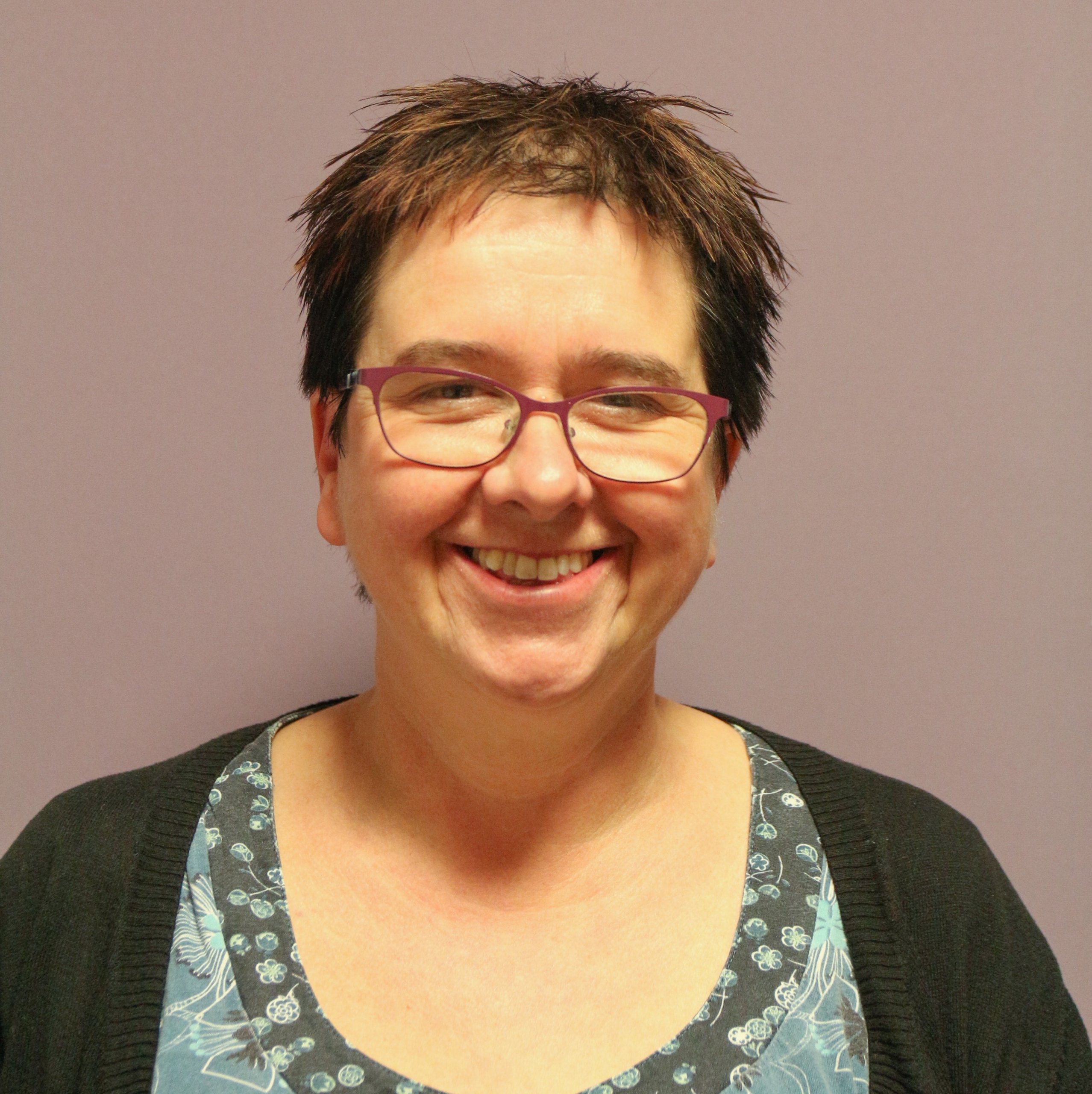 Jacquie has short, red-brown hair and glasses and is smiling in the photo.