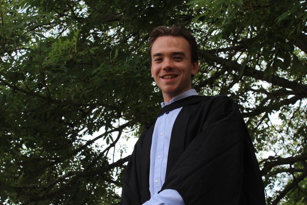 Morgan is pictured in graduation robes, standing beneath a tree.
