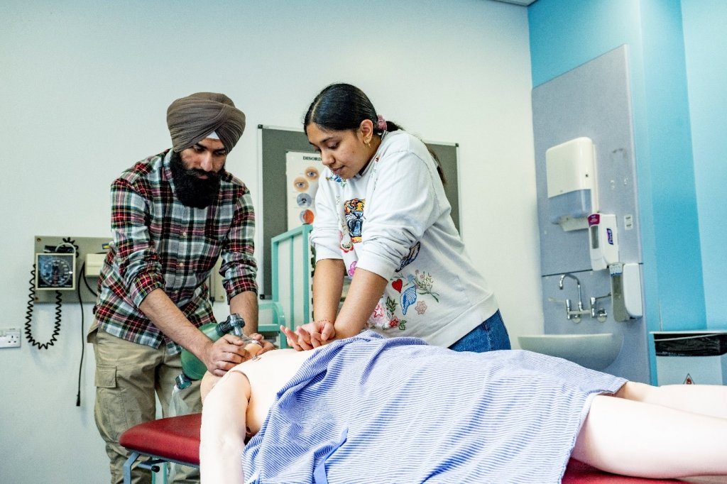 Two colleagues practice CPR on a dummy. The male colleague wears a turban and has a full beard, suggesting he is a practising Sikh.